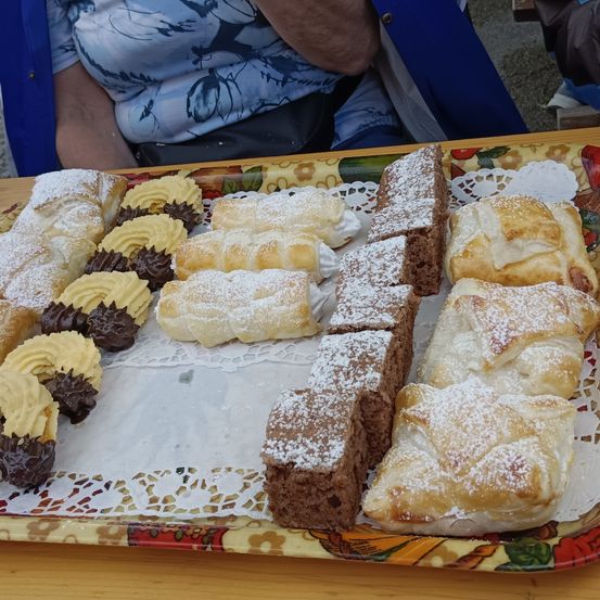 Auf einem Holztisch sind verschiedene Desserts zu sehen, darunter Croissants, Gebäck und Kuchen, die mit Puderzucker bestreut und auf einem Doily mit Blumenmuster platziert sind.