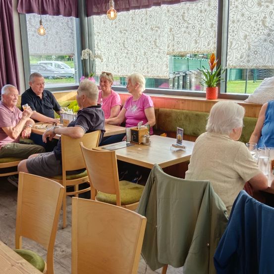 Eine Gruppe älterer Erwachsener sitzt in einem Café um einen Tisch herum und führt ein Gespräch. Der Tisch ist mit Gläsern und Essen gefüllt. Hinter ihnen befinden sich Fenster mit Blumenmustern.