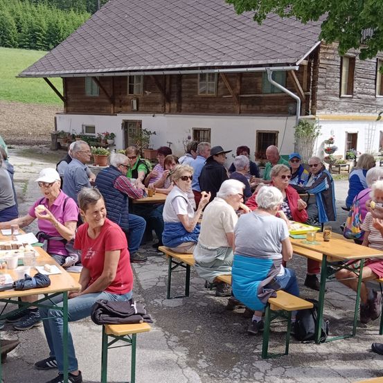 Eine Gruppe älterer Menschen sitzt auf Bänken vor einem Haus und isst. Einige tragen Hüte und Brillen. Die Tische sind mit Essen, Getränken und Servietten gedeckt. Das Haus hat einen Schornstein und Topfpflanzen.