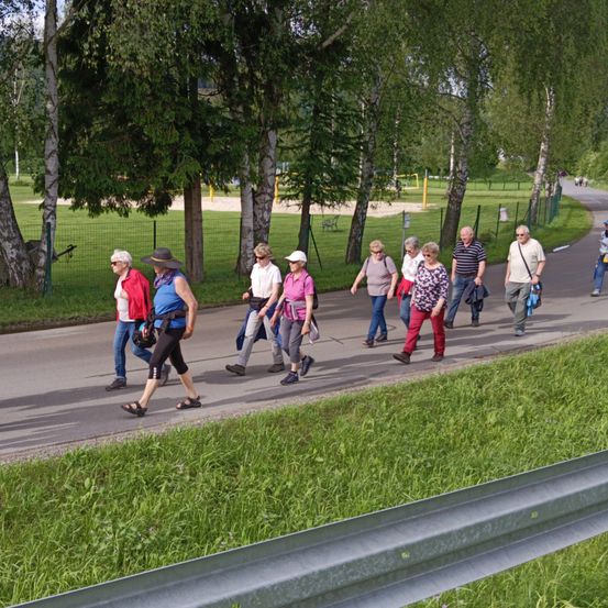 Eine Gruppe von Menschen läuft auf einem gepflasterten Weg, umgeben von Bäumen und Gras. Einige tragen Hüte und Sonnenbrillen, und eine trägt eine Handtasche. Der Weg ist mit einer Metallbarriere gesäumt.