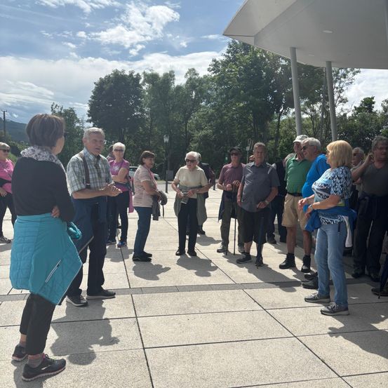 Eine Gruppe von Menschen steht draußen unter einem überdachten Bereich. Sie unterhalten sich, einige tragen Hüte und Sonnenbrillen. Bäume und ein bewölkter Himmel sind im Hintergrund.