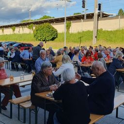 Ein Outdoor-Essbereich mit vielen älteren Menschen, die an Tischen sitzen, einige mit Brille und halten Gläser Bier. Im Hintergrund sind Bäume und eine Brücke.