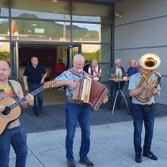 Eine Gruppe von Männern spielt Musikinstrumente vor einem Gebäude mit Glaswänden, die Berge überblicken. Einer spielt Gitarre, ein anderer Akkordeon und ein Dritter Tuba. Menschen stehen herum, einige beobachten.
