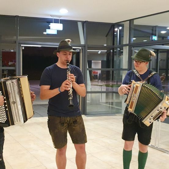 Drei Männer spielen in einem Gebäude Instrumente. Einer spielt Klarinette, ein anderer Akkordeon und der Dritte hält ein Akkordeon. Sie sind in traditioneller Kleidung gekleidet.