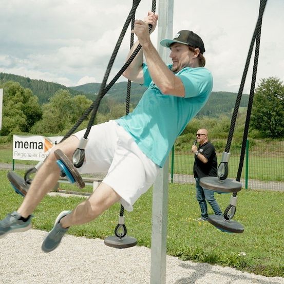 Ein Mann in einem türkisfarbenen T-Shirt und weißen Shorts hängt an einem sonnigen Tag an einem Schaukel. Ein anderer Mann steht in der Nähe, trägt eine Sonnenbrille und ein schwarzes T-Shirt.