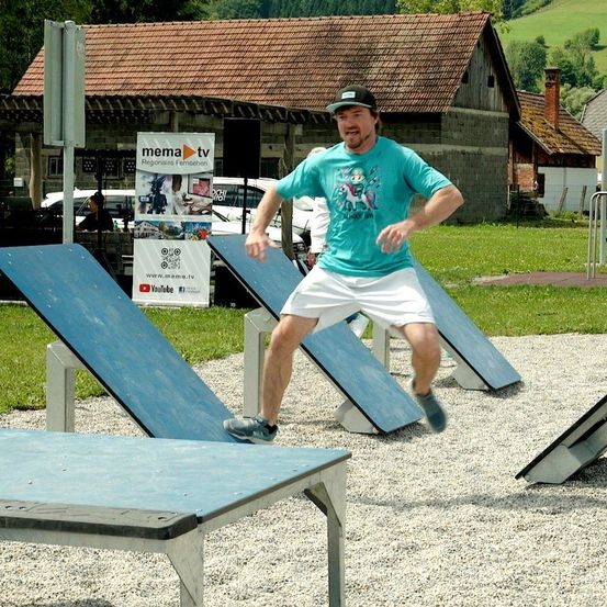 Ein Mann springt über Hindernisse in einem Schotterbereich. Er trägt ein türkisfarbenes T-Shirt und weiße Shorts. Hinter ihm stehen zwei Banner und ein Gebäude mit einem Schornstein.