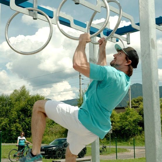 Ein Mann macht Klimmzüge an einer Outdoor-Übungsstange in einem Park. Er trägt ein blaues T-Shirt, weiße Shorts und eine Kappe. Hinter ihm fährt eine Frau mit dem Fahrrad. Im Hintergrund steht ein Auto und ein Berg.
