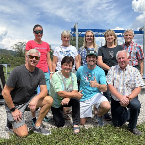 Eine Gruppe von Erwachsenen posiert für ein Foto auf einem Kiesweg, lächelt mit einem Spielplatz im Hintergrund unter einem teilweise bewölkten Himmel.