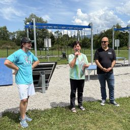 Drei Personen stehen im Freien in einem Park. Eine Frau hält ein Mikrofon, während ein Mann neben ihr und ein anderer Mann hinter ihr steht. Im Hintergrund befinden sich Übungsgeräte und ein Zaun.