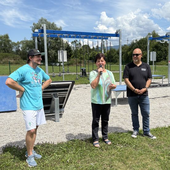 Drei Personen stehen im Freien in einem Park. Eine Frau hält ein Mikrofon, während ein Mann neben ihr und ein anderer Mann hinter ihr steht. Im Hintergrund befinden sich Übungsgeräte und ein Zaun.