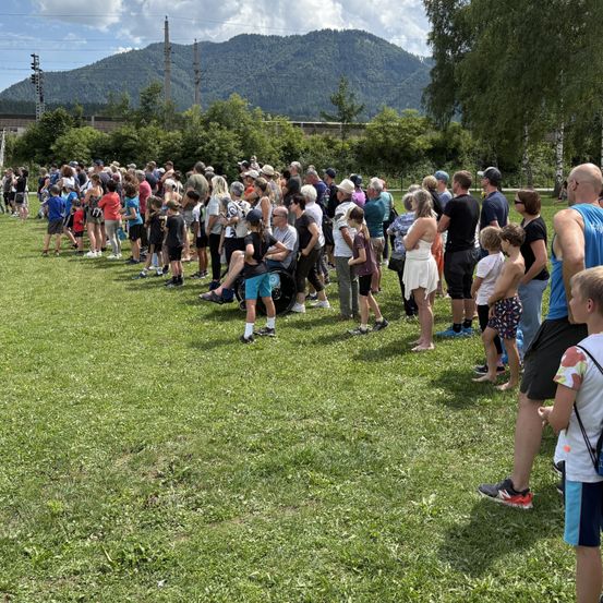 Eine große Menschenmenge versammelt sich auf einem Rasenfeld mit Bergen im Hintergrund. Sie stehen in einer Reihe, einige sitzen auf dem Boden, unter einem teilweise bewölkten Himmel.