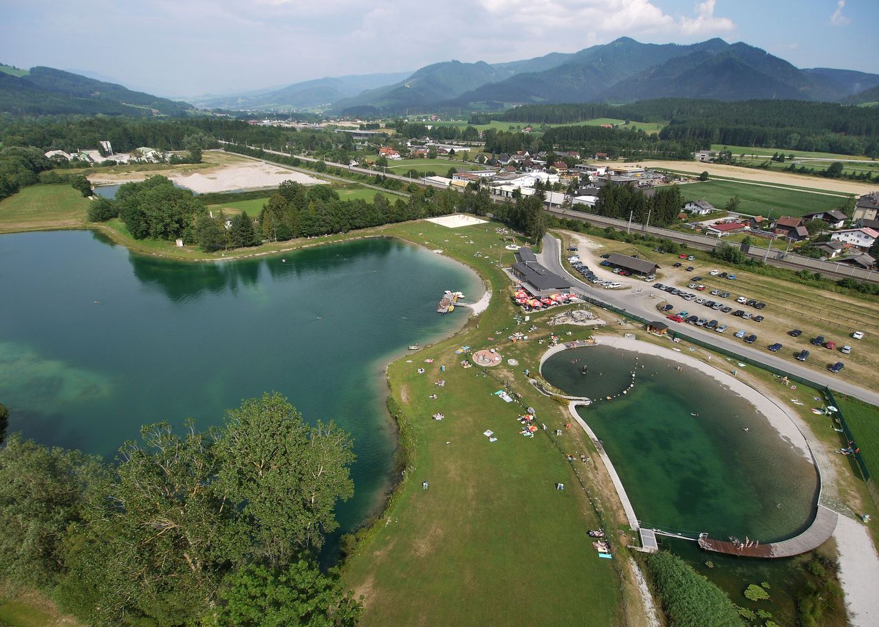 Ein Luftbild eines friedlichen Sees mit schwimmenden, sonnenbadenden und auf der Wiese sitzenden Menschen. In der Nähe befinden sich Gebäude, eine Straße und geparkte Autos. Der See ist von üppigem Grün und Bergen in der Ferne umgeben.