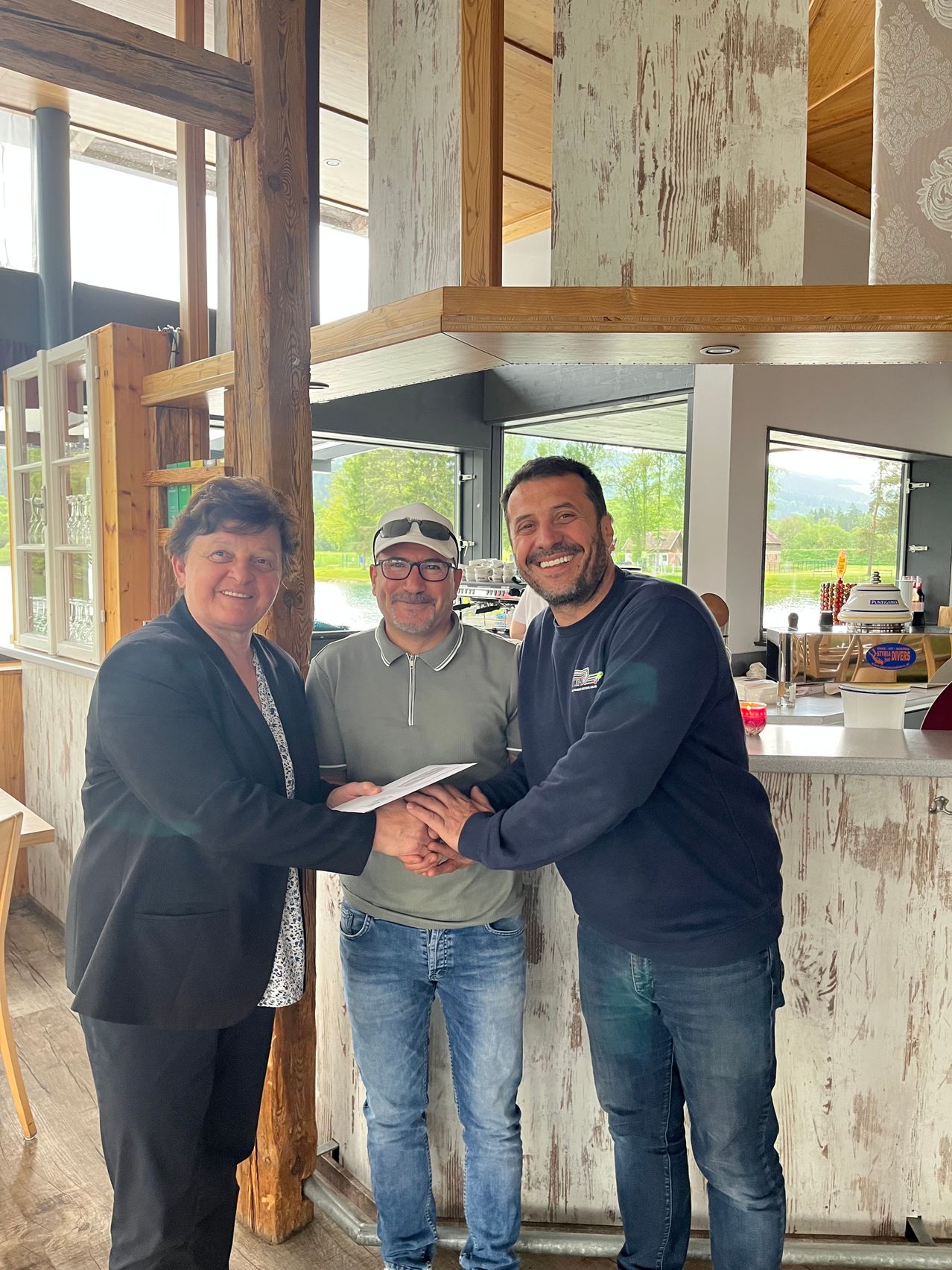 Drei Personen posieren für ein Foto in einem Restaurant. Der Mann in der Mitte lächelt und hält ein Stück Papier. Die anderen beiden lächeln ebenfalls. Hinter ihnen ist eine Theke mit einigen Gegenständen darauf.