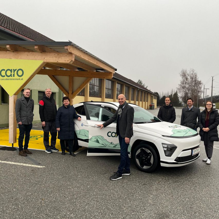 Eine Gruppe von Menschen in Winterkleidung steht vor einem weißen Elektroauto mit Logo, das an einer Ladestation geparkt ist.
