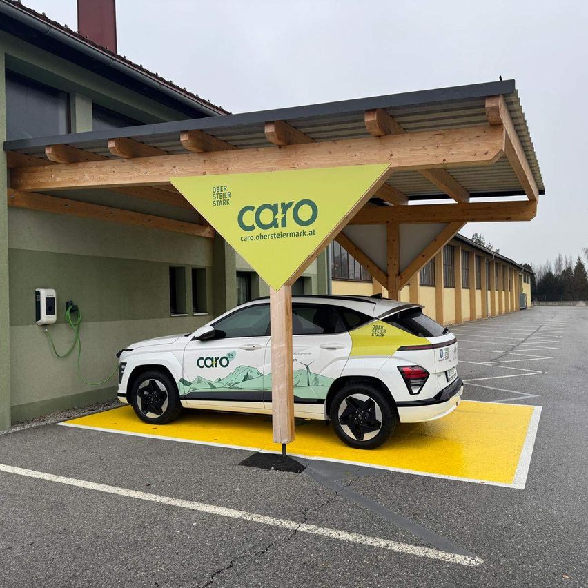 Ein weißer Caro SUV ist unter einem hölzernen Vordach mit einem gelben Schild geparkt, neben einer Ladestation. Der SUV steht auf einer gelben Plattform in einem Parkplatz.