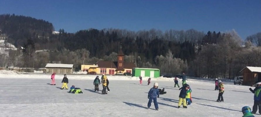 Eine Gruppe von Kindern spielt auf einem verschneiten Feld. Sie tragen Winterkleidung und einige skaten. Im Hintergrund steht ein Gebäude und ein Berg mit Bäumen.