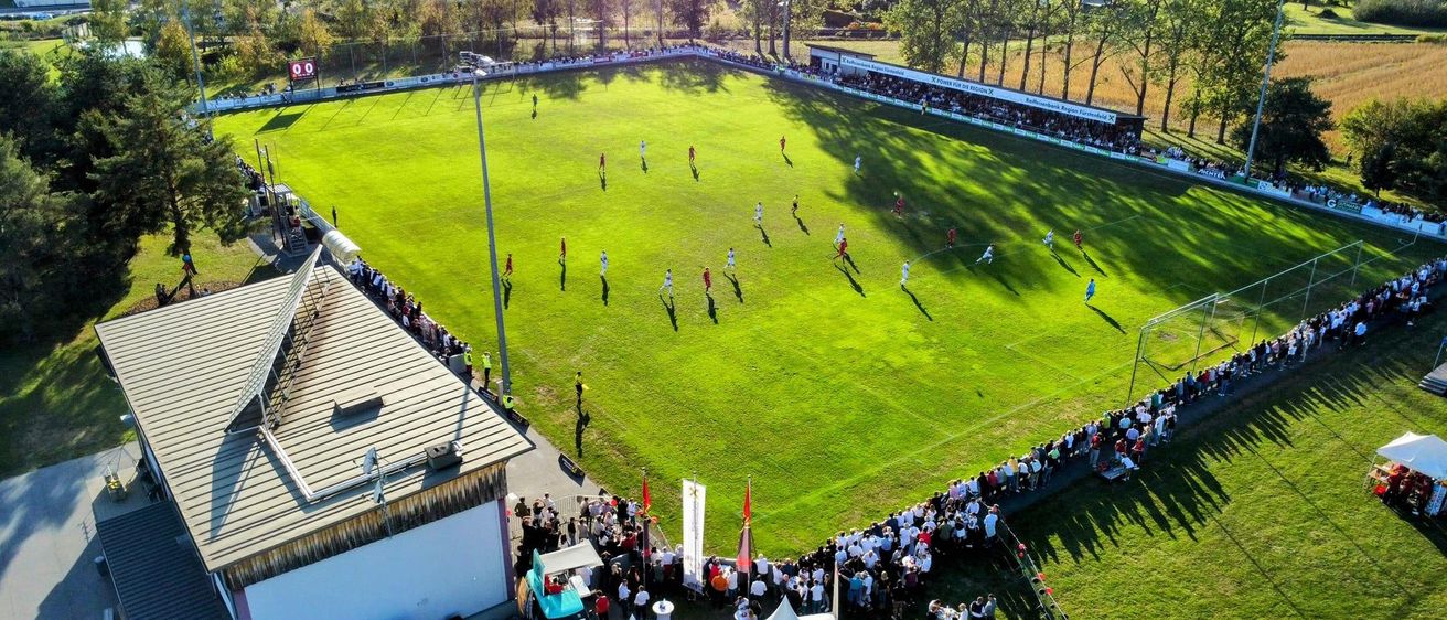 Eine Luftaufnahme eines Fußballfeldes mit Spielern in roten und weißen Uniformen. Das Feld ist von einer großen Menge von Zuschauern und Zelten umgeben.