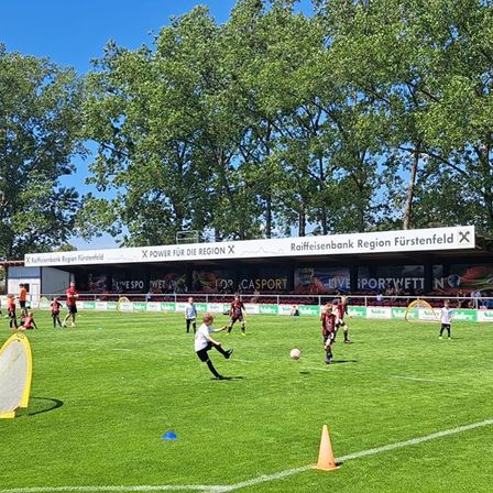 Kinder spielen Fussball auf einem Feld mit einem Banner, das Raiffeisenbank Region Furstenfeld im Hintergrund bewirbt.