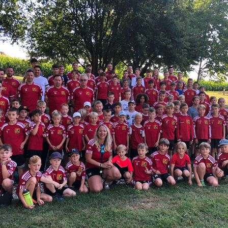 Eine Fußballmannschaft mit Kindern und Erwachsenen in roten Trikots steht auf einem Feld mit einem Baum im Hintergrund.