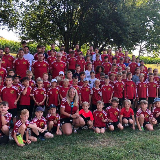 Eine Fußballmannschaft mit Kindern und Erwachsenen in roten Trikots steht auf einem Feld mit einem Baum im Hintergrund.