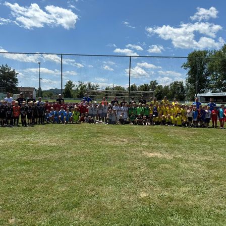 Eine Gruppe von Kindern und Erwachsenen steht auf einem Feld vor einem Zaun, mit einem Fußballtor im Hintergrund. Sie tragen verschiedenfarbige Trikots und einige haben Medaillen.