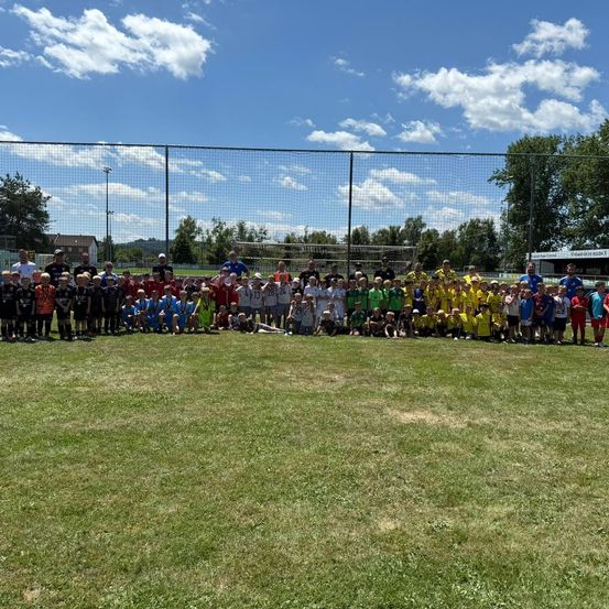 Eine Gruppe von Kindern und Erwachsenen steht auf einem Feld vor einem Zaun, mit einem Fußballtor im Hintergrund. Sie tragen verschiedenfarbige Trikots und einige haben Medaillen.