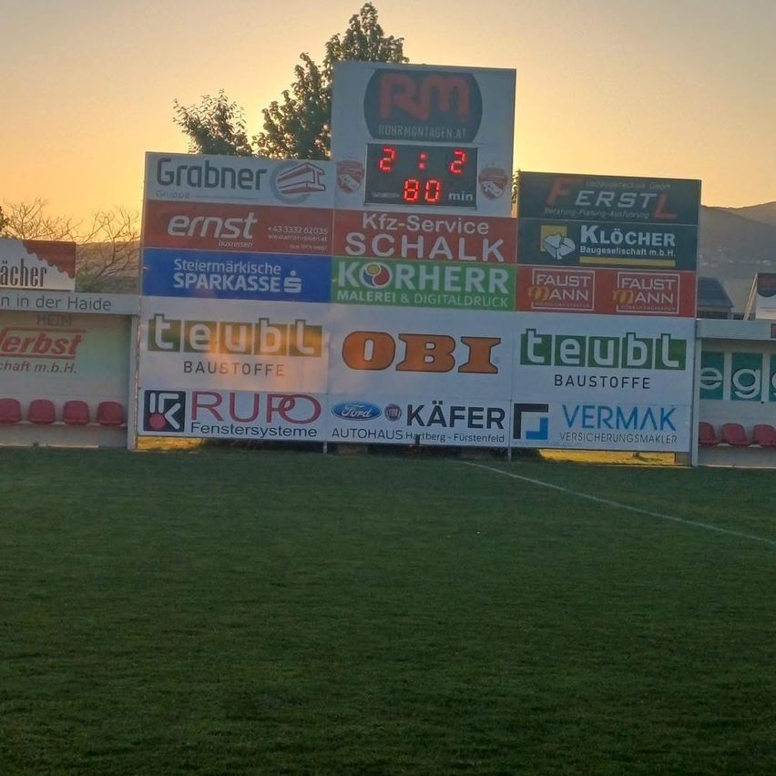 Ein Sportplatz mit Anzeigetafel und verschiedenen Werbebannern. Die Anzeigetafel zeigt einen Stand von 2:2 bei 80 gespielten Minuten. Es gibt Werbung für Grabner, Kfz-Service usw.