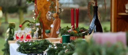 Ein festlicher Tisch, geschmückt mit Tannengirlanden, roten Kerzen und einer dekorierten Flasche. Eine Holzstatue und andere Weihnachtsdekorationen im Hintergrund.