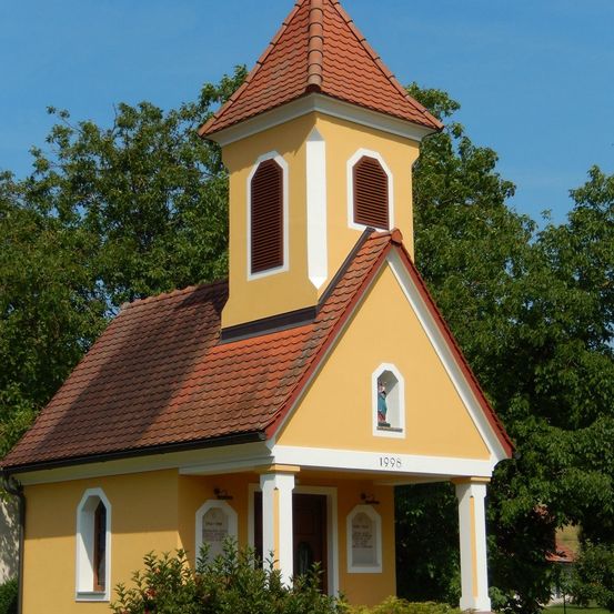 Eine kleine gelbe Kapelle mit rotem Dach, erbaut im Jahr 1998, steht vor einem Hintergrund üppiger grüner Bäume.