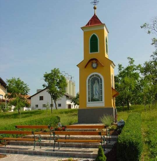 Eine kleine gelbe Kapelle mit rotem Dach und einer Statue der Maria steht in einem Grasfeld mit Bänken. Bäume und ein weißes Haus sind im Hintergrund.