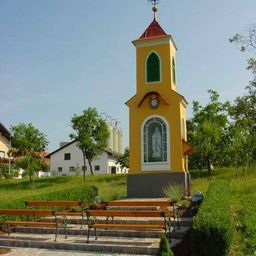 Eine kleine gelbe Kapelle mit rotem Dach und einer Statue der Maria steht in einem Grasfeld mit Bänken. Bäume und ein weißes Haus sind im Hintergrund.