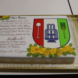 Ein Kuchen mit einer Ruhestandsnachricht, einem Schild mit einem Logo und Blumen auf weißem Hintergrund. Die Nachricht ist auf Deutsch geschrieben.