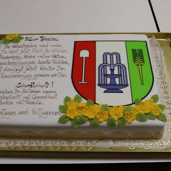 Ein Kuchen mit einer Ruhestandsnachricht, einem Schild mit einem Logo und Blumen auf weißem Hintergrund. Die Nachricht ist auf Deutsch geschrieben.