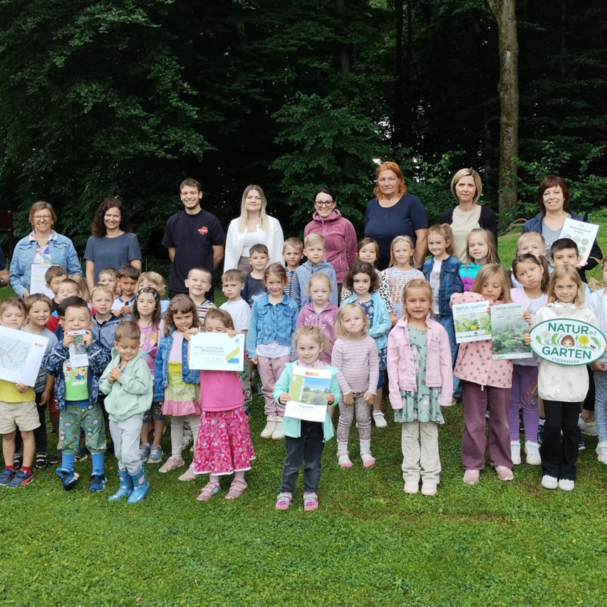 Eine Gruppenaufnahme von Kindern und Erwachsenen, die in einem Park mit grünen Schildern stehen, Urkunden halten und für ein Foto posieren.