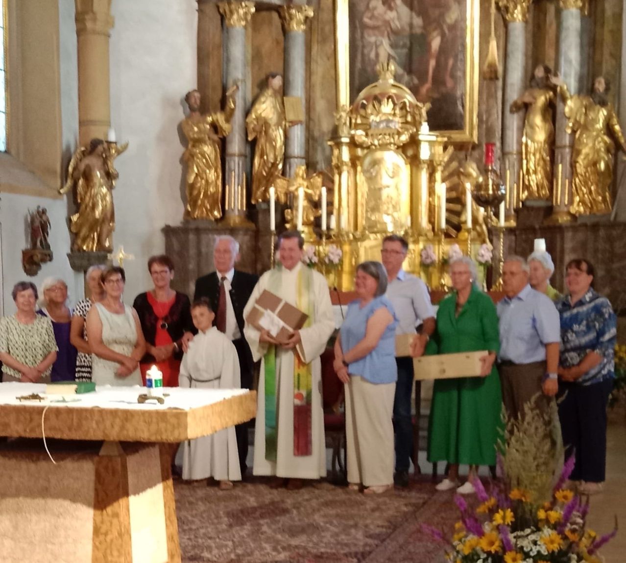 Ein Priester hält ein Buch in einer Kirche mit einem Altar, Kerzen und Statuen. Vor ihm stehen Menschen, darunter ein Kind. Blumen und Kerzen stehen auf dem Altar.
