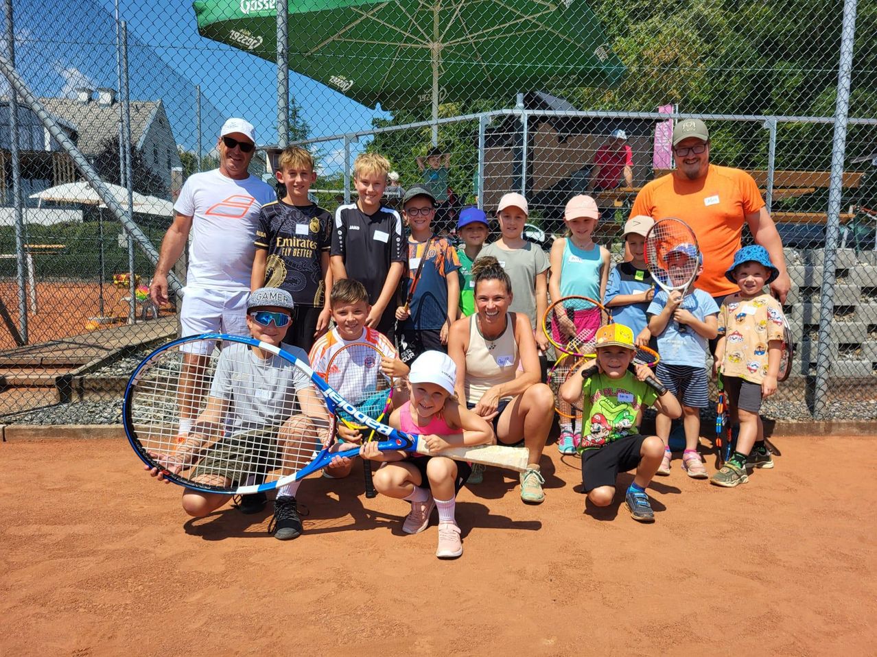 Eine Gruppe von Kindern und Erwachsenen posiert für ein Foto auf einem Tennisplatz mit Tennisschlägern in der Hand.