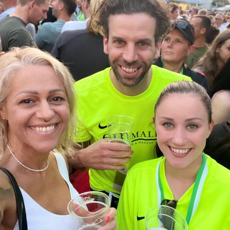 Drei Personen lächeln in die Kamera, wahrscheinlich bei einem Festival. Ein Mann hält einen Becher, und zwei Frauen stehen auf beiden Seiten. Der Hintergrund ist unscharf mit vielen Leuten.