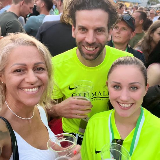 Drei Personen lächeln in die Kamera, wahrscheinlich bei einem Festival. Ein Mann hält einen Becher, und zwei Frauen stehen auf beiden Seiten. Der Hintergrund ist unscharf mit vielen Leuten.