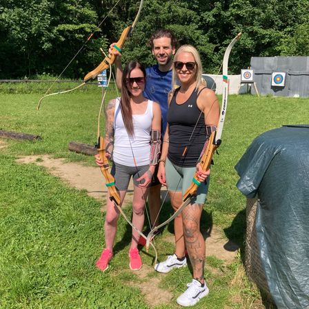 Drei Personen, zwei Frauen und ein Mann, stehen mit Bogen in einer grasigen Freifläche. Hinter ihnen befinden sich Holzziele und ein Holzzaun.