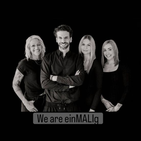 Ein schwarz-weißes Bild von vier Menschen, die zusammenstehen und lächeln, wobei der Mann in der Mitte steht und unten die Worte 'Wir sind einMALig' stehen.