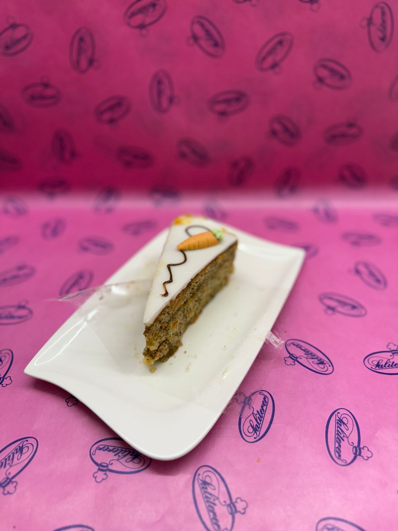 Ein Stück Karottenkuchen mit weißer Glasur und einer Karotte darauf steht auf einem weißen Teller auf einer rosa Oberfläche.