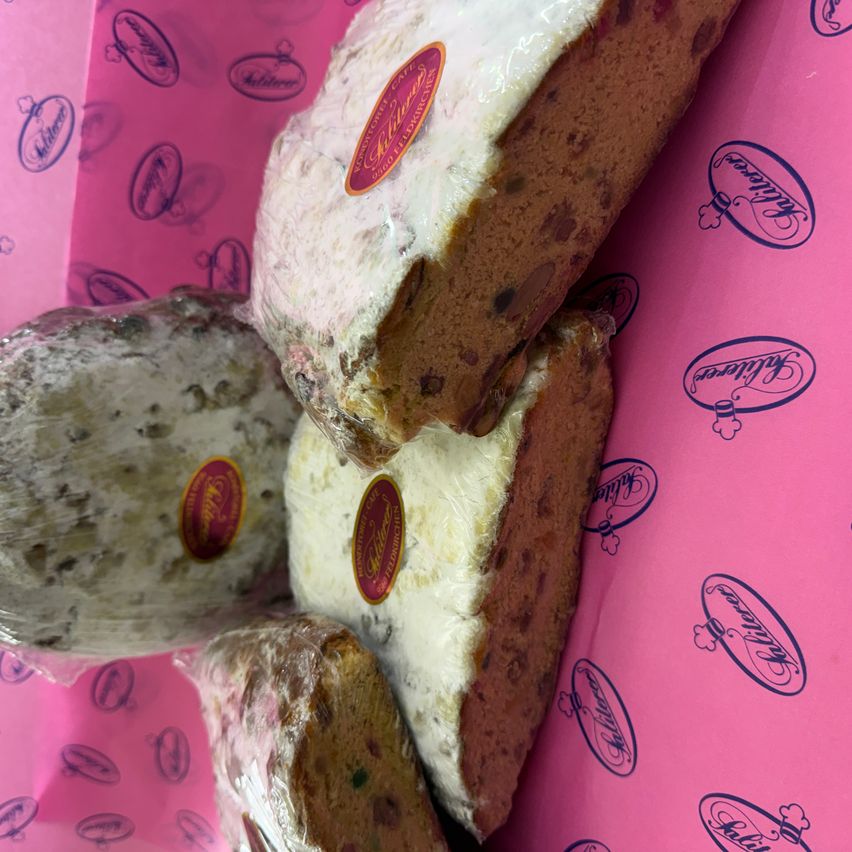 Mehrere Scheiben Fruchtkuchen sind in Plastik eingewickelt und auf einem rosa Hintergrund angeordnet. Jede Scheibe hat ein rotes Etikett mit Text und Logo. Der Fruchtkuchen enthält getrocknete Früchte und Nüsse.