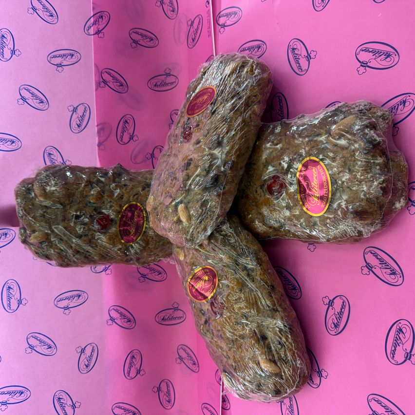 Drei einzeln verpackte Stücke Fruchtkuchen, jedes mit einem roten Etikett, sind in einem X-förmigen Muster auf einer rosa Oberfläche mit blauen Logos platziert.