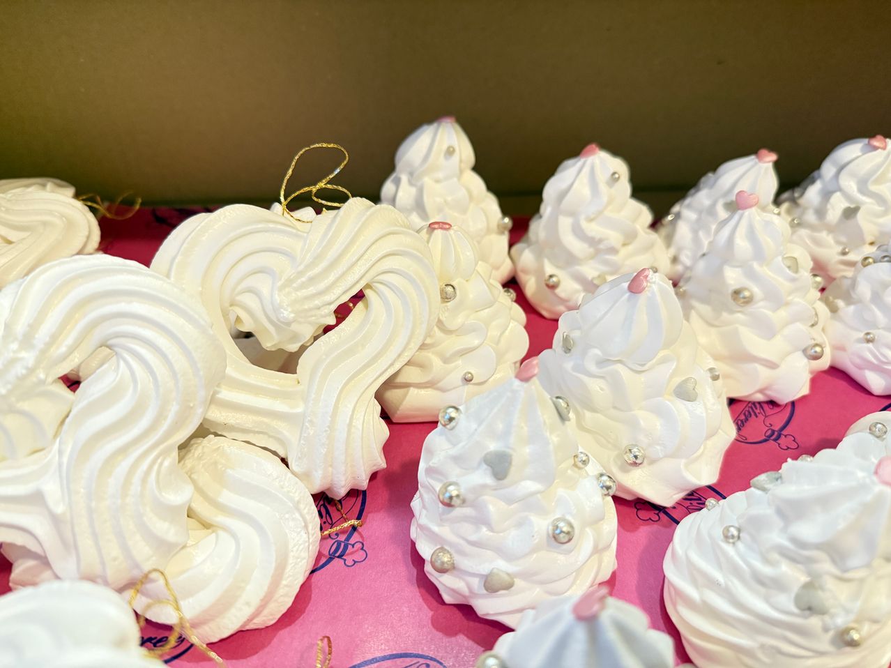 Mehrere weiße Baiser in Form von Weihnachtsbäumen mit goldenen und silbernen Perlen sind auf einer rosa Oberfläche ausgestellt. Die Baiser haben komplizierte Designs.