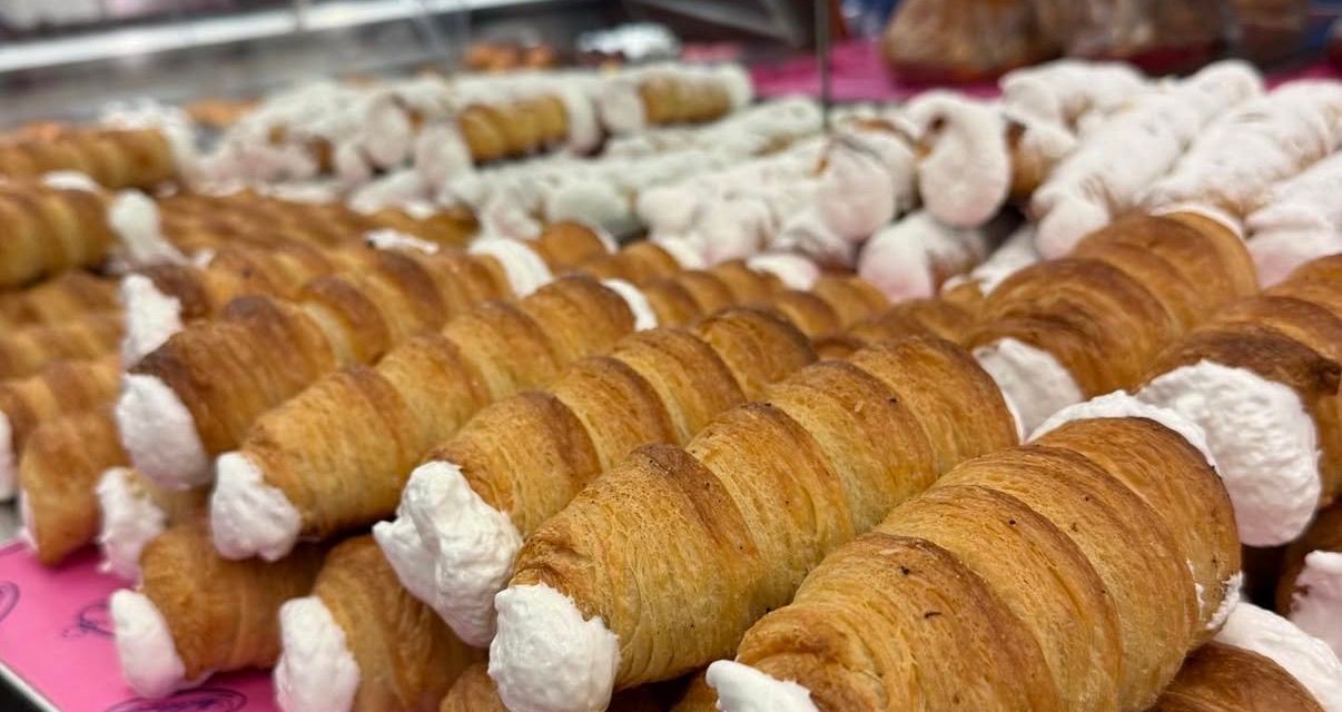 A display of multiple croissants with cream on top, some with visible spiral patterns, arranged in a row on a pink surface.