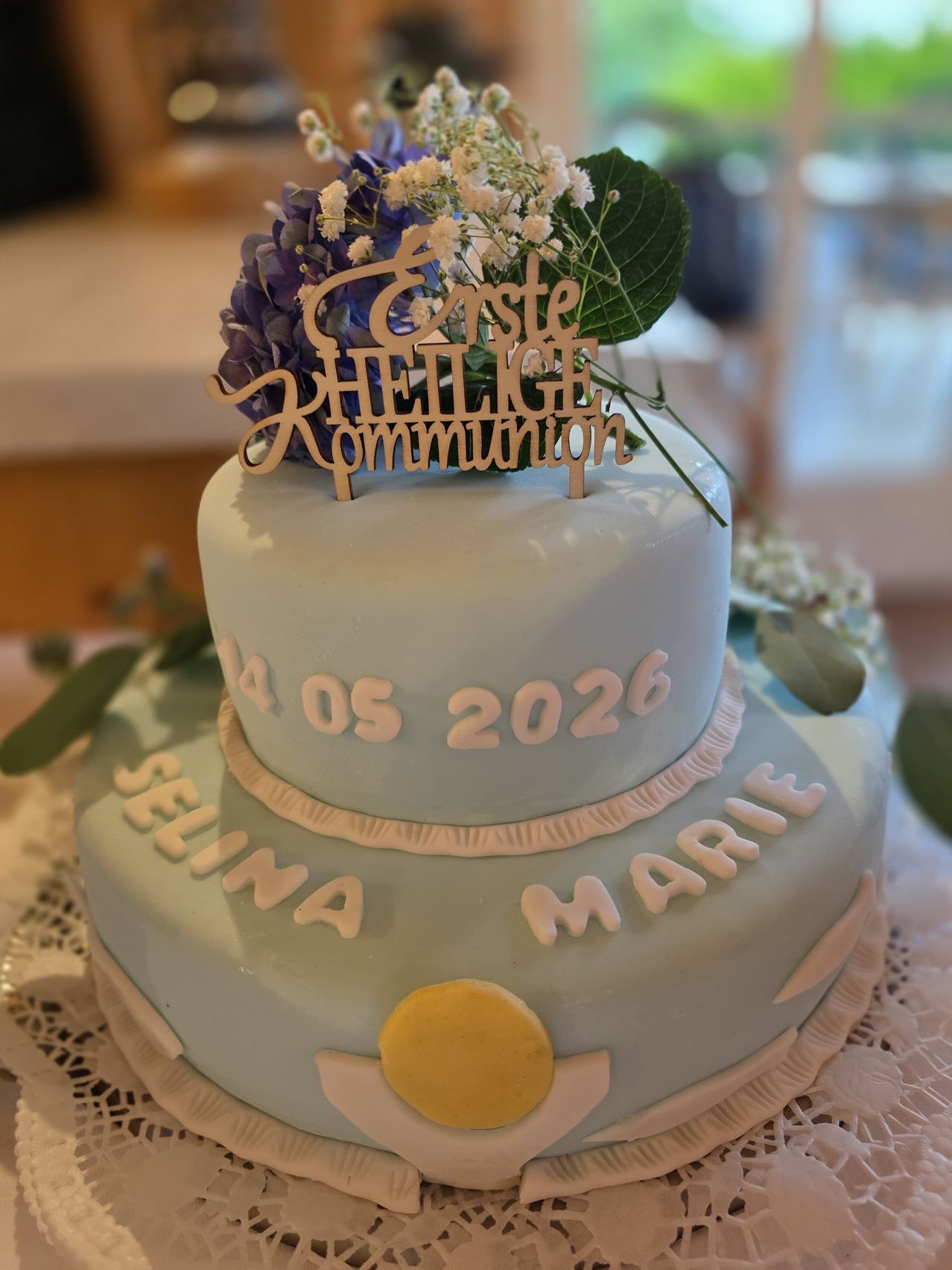 Ein zweistöckiger Kuchen mit blauer Glasur, weißen Blumen und grünen Blättern. Es steht 'erste HEILIGE Kommunion' und 'SELINA MARIE 14.05.2026'. Der Kuchen steht auf einem Spitzendeckchen.