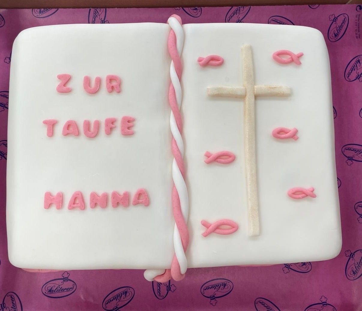 Ein Buch geformter Kuchen mit rosa und weißen Dekorationen. Es steht 'zur Taufe' und 'Mamma' darauf. Es gibt auch rosa Kreuze und Fischdesigns.