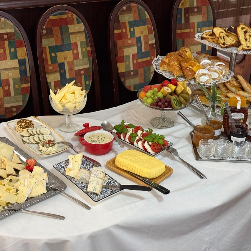 Ein Tisch ist mit einem weißen Tuch bedeckt und zeigt eine Auswahl an Käse, Obst und Gebäck. Verschiedene Käsesorten sind auf Tabletts angeordnet. Schalen mit Obst und Brot stehen auf einem mehrstöckigen Ständer.