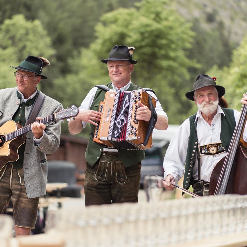 Drei Männer in traditioneller deutscher Kleidung spielen Musik im Freien. Einer spielt Gitarre, ein anderer Akkordeon und der dritte Cello. Sie sind von Gläsern und einer landschaftlichen Kulisse aus Bäumen umgeben.