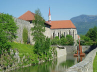 Ein historisches Gebäude mit einem roten Dach und einem Turm steht neben einem Fluss. Dichtes Grün und Bäume umgeben das Gebäude, und Berge sind im Hintergrund sichtbar.
